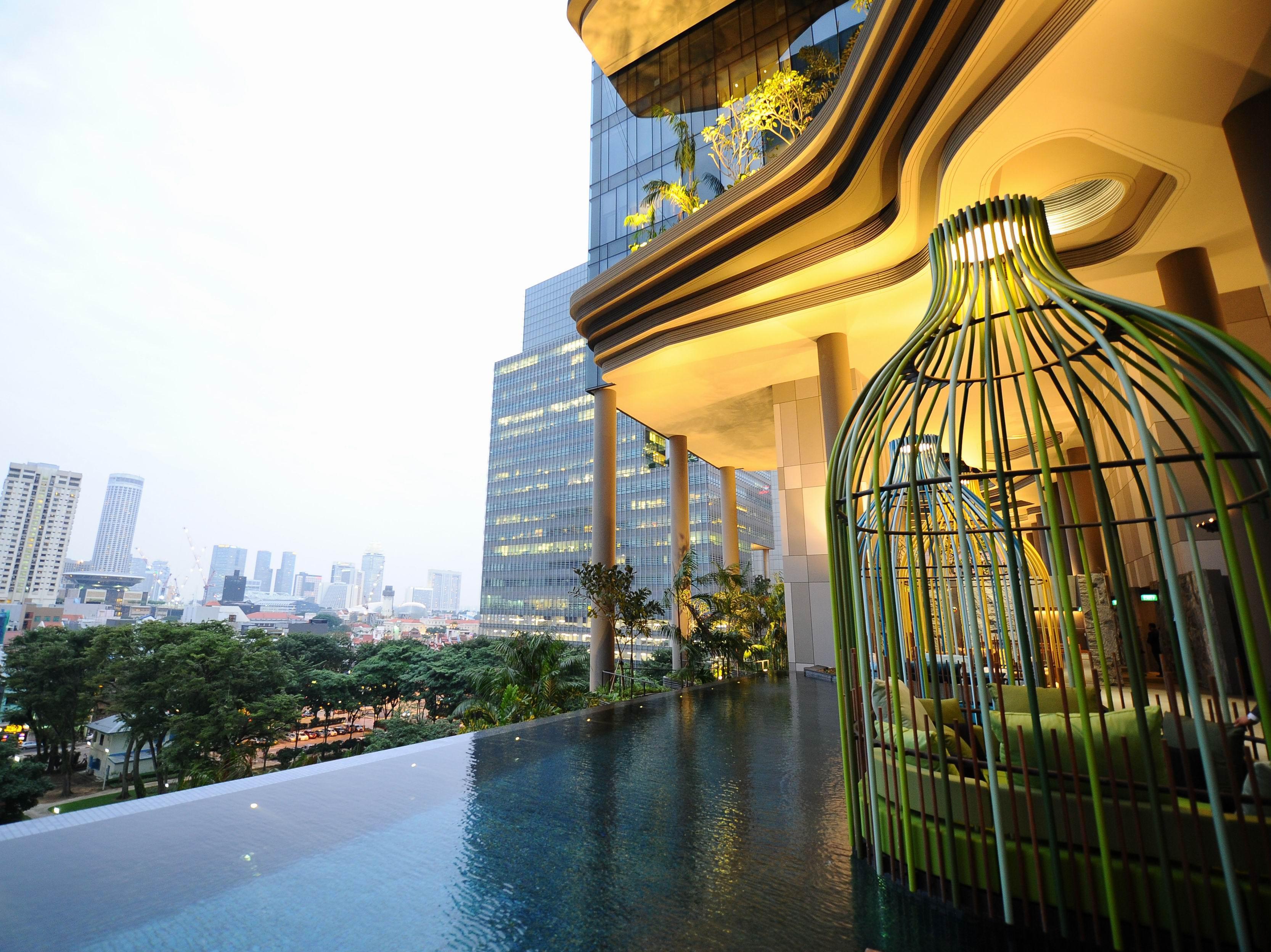 新加坡皮克林宾乐雅臻选酒店 外观 照片 The photo showcases a modern, luxurious space that features an infinity pool with a sleek, reflective surface. In the foreground, there is a unique structure resembling a large birdcage, designed with green elements. The background includes tall city