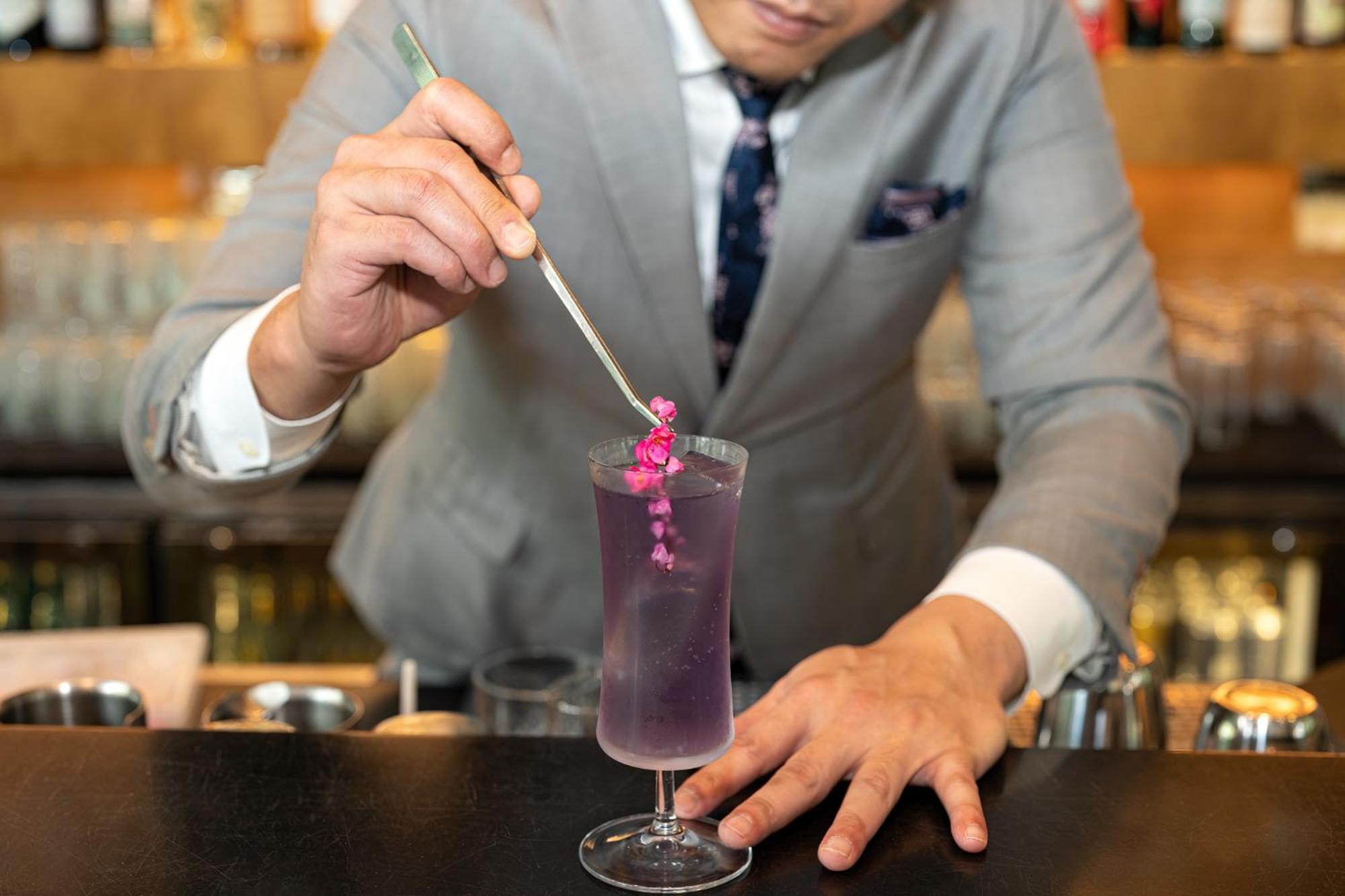 新加坡皮克林宾乐雅臻选酒店 外观 照片 The photo shows a bartender in a stylish gray suit preparing a cocktail. He is holding a long, thin tool, possibly a stirrer or a tweezer, and is carefully placing a small pink flower garnish into a tall glass filled with a vibrant purple drink. The 