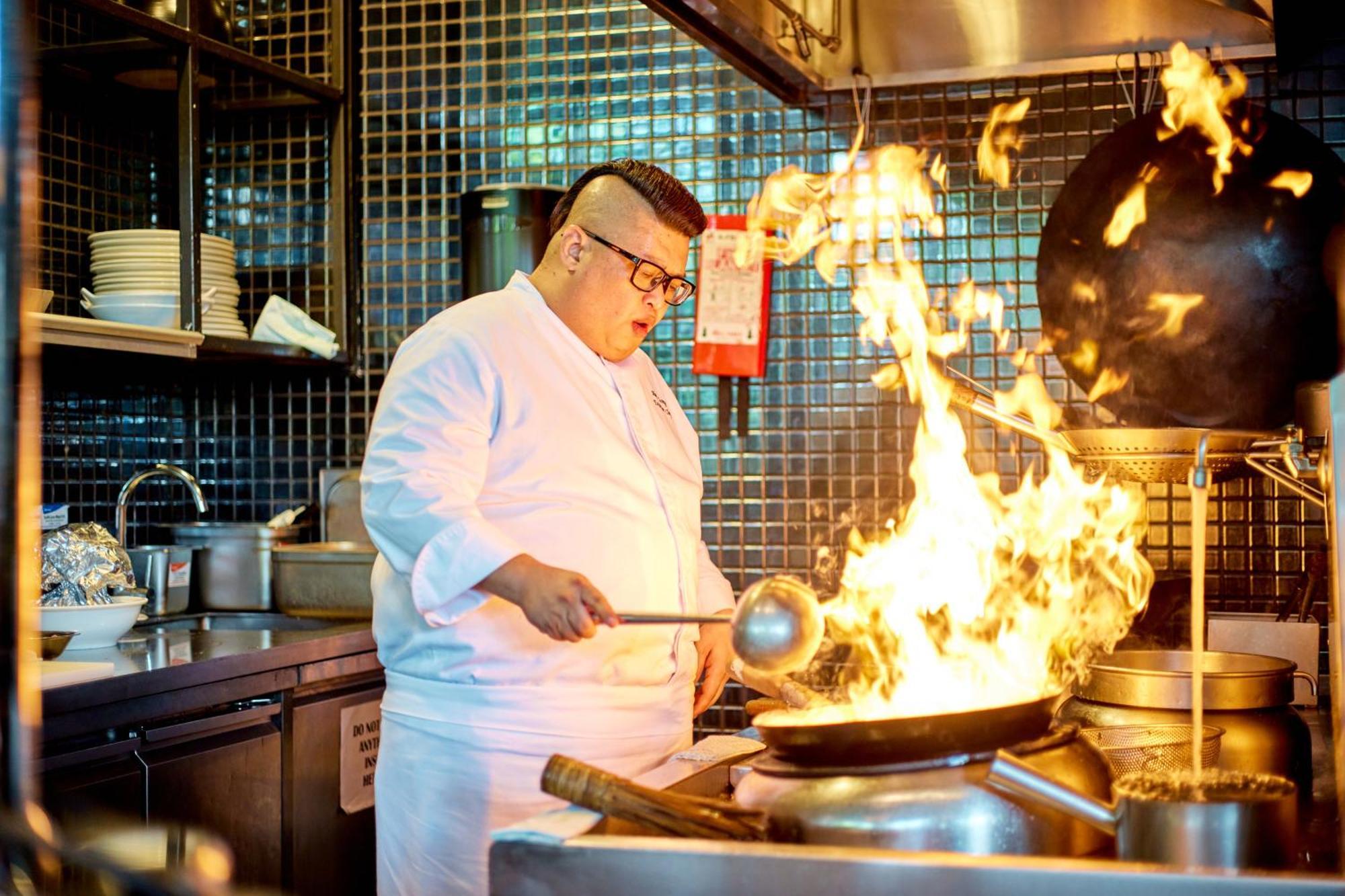 新加坡皮克林宾乐雅臻选酒店 外观 照片 The photo shows a chef in a kitchen, wearing a white chef's jacket and glasses. He is actively cooking in front of a large wok that is producing a dramatic surge of flames. The kitchen has a modern look, with tiled walls and a variety of kitchen equi