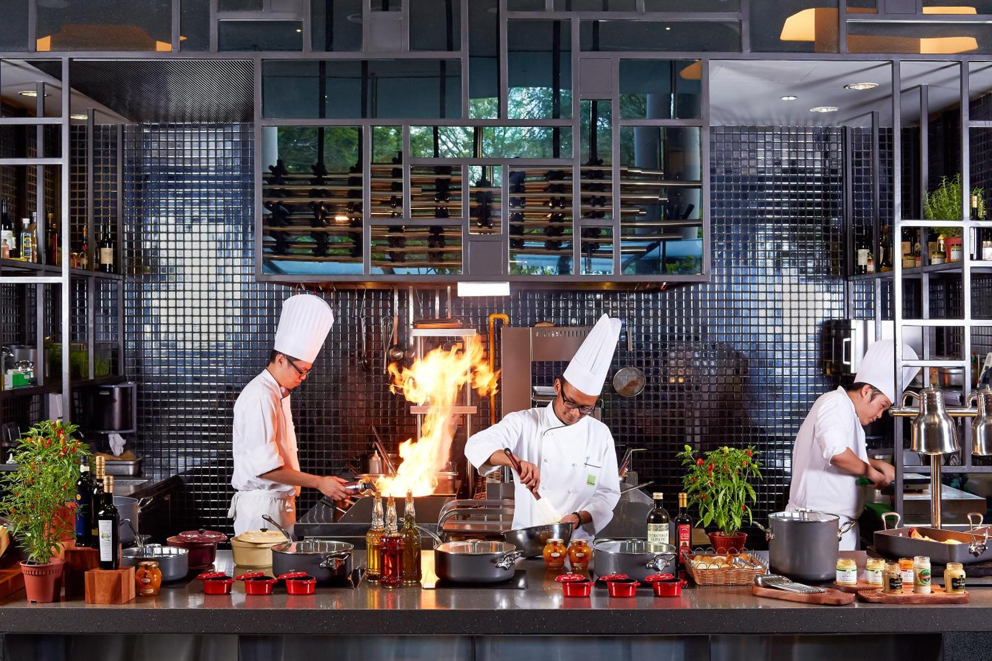 新加坡皮克林宾乐雅臻选酒店 外观 照片 The photo depicts a modern kitchen where three chefs are actively engaged in cooking. Two of the chefs appear to be stirring pots on the stove, and one chef is working with a flame, likely performing a technique that involves flambéing. The kitchen i