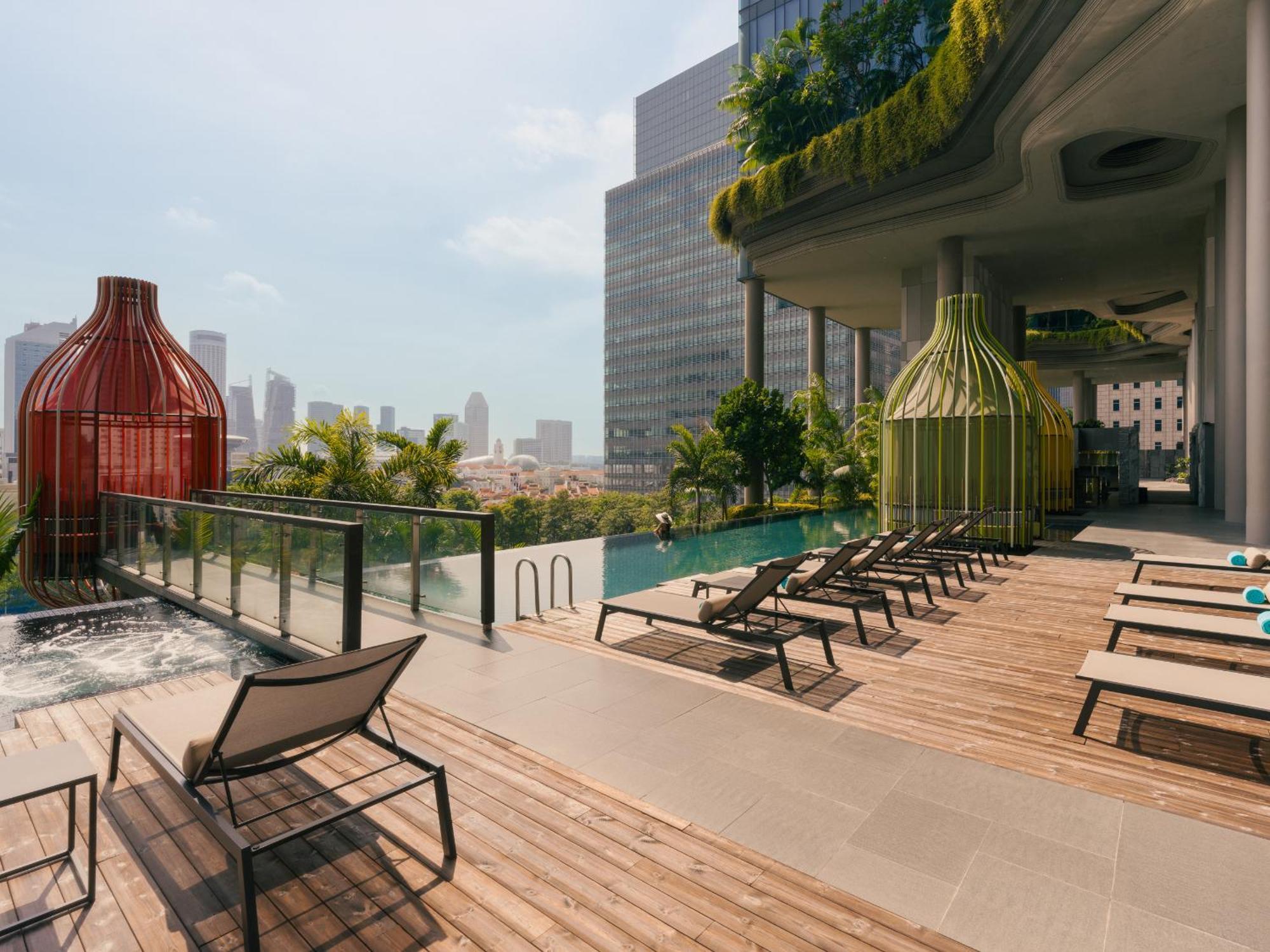 新加坡皮克林宾乐雅臻选酒店 外观 照片 The photo depicts a modern outdoor pool area of a high-rise building. There are lounge chairs arranged along a wooden deck beside a pool. Two large, sculptural structures in vibrant red and green colors are prominently placed near the pool, likely se